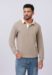 Beige long-sleeve polo shirt with a cream collar, featuring a small yellow logo on the chest. Paired with light blue jeans.