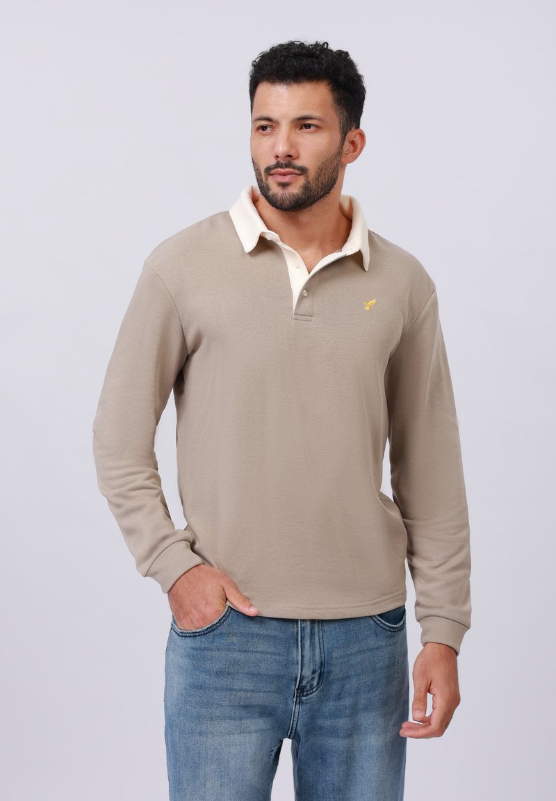 Beige long-sleeve polo shirt with a cream collar, featuring a small yellow logo on the chest. Paired with light blue jeans.