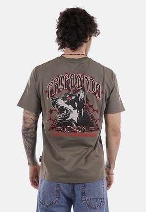 Uomo con capelli ricci visto di spalle indossa una maglietta marrone con un cane ringhiante, catene e il testo "PROPAGANDA ENTRA A TUO RISCHIO".