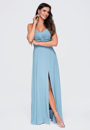 Mujer con vestido azul claro sin mangas con una abertura en el muslo, lleva tacones plateados y grandes pendientes florales, sonriendo y posando.