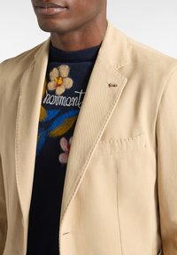 Beige blazer i slätt tyg, med en enkel ficka och en pin-detalj. Bärs över en marinblå tröja med blommig brodyr.