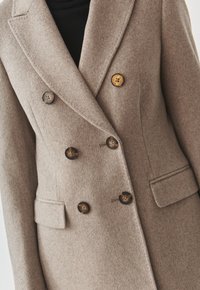Abrigo beige de doble botonadura hecho de lana, con solapas muesca, dos bolsillos frontales y cuatro botones oscuros. Textura suave y corte entallado.