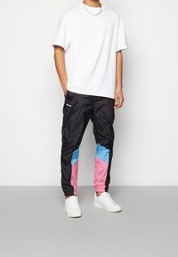 Man wearing white sneakers, black track pants with blue and pink color blocks, and a plain white T-shirt, standing with one hand in pocket.