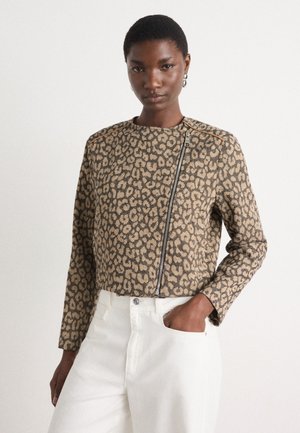 Veste courte en imprimé léopard beige et noir, dotée d'une fermeture éclair et d'épaules structurées, associée à un pantalon blanc.