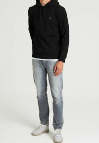 Felpa con cappuccio nera con coulisse regolabili e piccolo logo sul petto, abbinata a jeans azzurri e sneaker grigie.