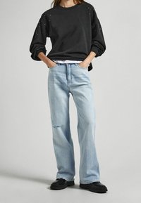Persona vistiendo un sudadera negra con pequeños puntos, jeans rectos rasgados de color azul claro, zapatos negros, de pie con las manos en los bolsillos.