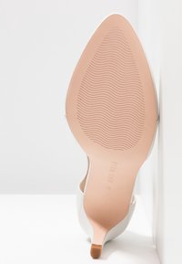 Chaussure blanche à talon haut avec une pointe pointue, un extérieur lisse et une semelle rose texturée présentant des lignes ondulées. Marque claire visible.
