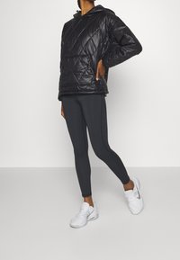 Pullover nero, imbottito, con cappuccio e tasca frontale, abbinato a leggings aderenti grigio scuro e scarpe da ginnastica bianche.