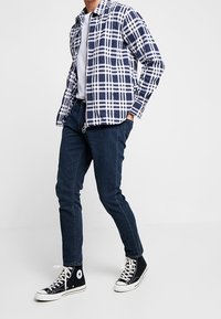 Rutigt marinblått och vitt zip-up skjorta, mörkblå slim-fit jeans och svarta högtoppsneakers med vita sulor. Avslappnad, lager-stil.