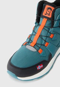 Teal stoff fottur med oransje snorer, grå detaljer, svart såle og et lite flaggmerke. Har polstret krage og teksturert materiale.