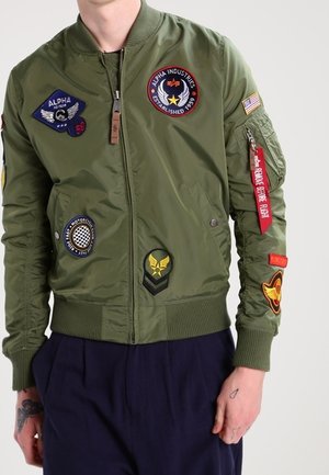 Olivgrön bomberjacka med märken på bröstet och ärmarna, ribbade cuffs och fåll, dragkedja och en slät, blank yta.