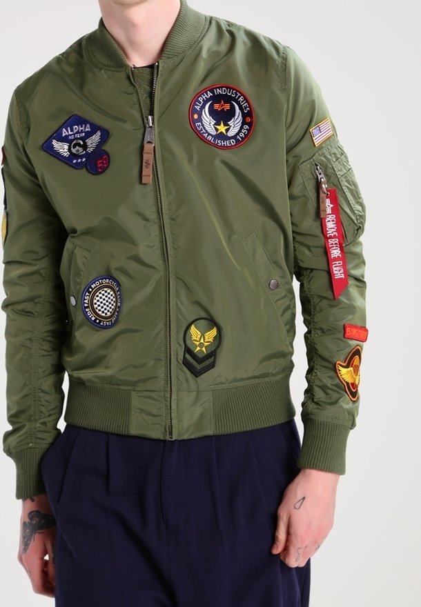 Giubbotto bomber verde oliva con distintivi sul petto e sulle maniche, polsini e orlo a coste, chiusura con zip e texture liscia e lucida.