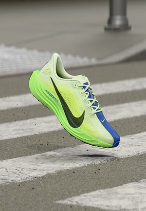 Neon groene en blauwe hardloopschoen met zwart logo, in de lucht zwevend boven een zebrapad op een asfaltweg.