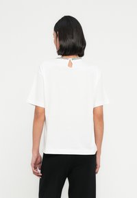 Λευκό βαμβακερό μπλουζάκι με κοντά μανίκια, στρογγυλή λαιμόκοψη με άνοιγμα με κλειδαρότρυπα στο πίσω μέρος και διακοσμητικές ασημένιες λεπτομέρειες στη λαιμόκοψη.