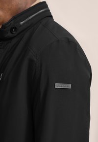 Schwarze Jacke mit hohem Kragen, glatter Textur und einem Metall-Logo auf dem Ärmel. Detaillierte Nähte sind entlang der Säume sichtbar.