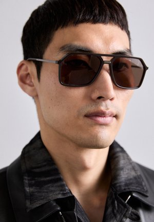 Brūnas pārspīlētas saulesbrilles ar ģeometrisku formu un plānu melnu metāla rāmi, valkātas kopā ar tumšu jakas ar apkakli, kurai ir teksturēts raksts.