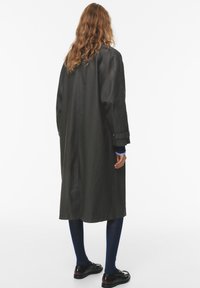 Cappotto lungo di un grigio scuro con taglio dritto, colletto a rever e polsini con bottoni. Indossato sopra collant blu e scarpe nere. Tessuto dalla texture liscia.