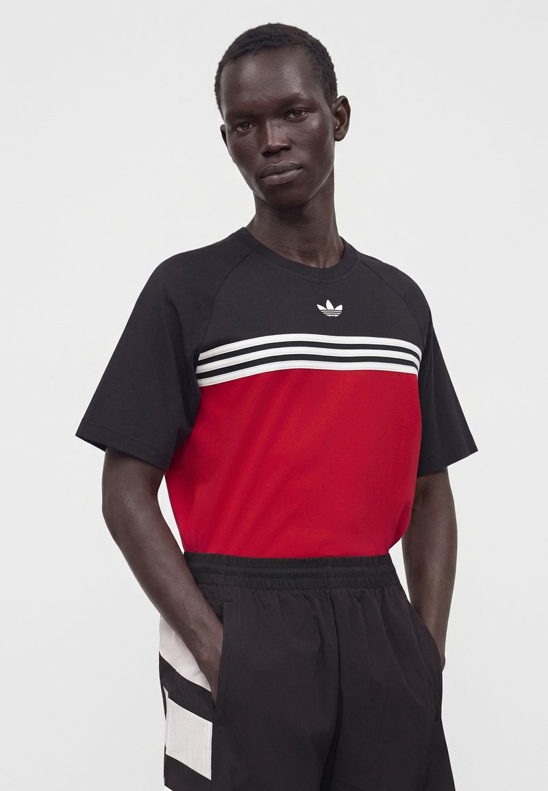 Mand iført en sort og rød Adidas t-shirt med hvide striber, sorte træningsbukser med hvide sidelommer, står med hænderne i lommerne.