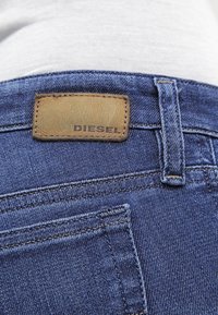 Primer plano de un bolsillo trasero de jeans de mezclilla azul y un lazo para cinturón con una etiqueta de cuero marrón de la marca Diesel sobre el bolsillo.