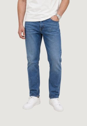 Mand iført blå jeans, hvide sneakers og en hvid t-shirt, stående med den ene hånd i lommen og den anden afslappet ved siden.