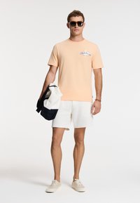 Lyse orange bomulds t-shirt med blåt tekstdesign, parret med hvide shorts. Holder en cremefarvet og marineblå taske, bærer solbriller og lyse sneakers.