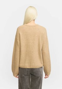 Beige übergroßer Pullover aus weichmaterial, mit Rundhalsausschnitt, langen Ärmeln und geripptem Saum. Getragen mit dunkelgrauten Hosen.