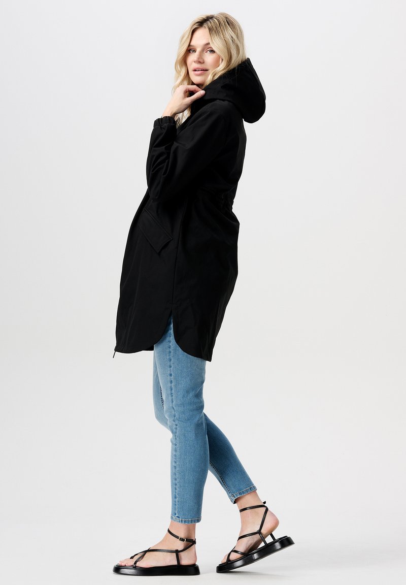 Schwarze, wasserdichte Jacke mit Kapuze, elastischem Bund und zwei Seitentaschen. Das Modell trägt hellblaue enge Jeans und schwarze Sandalen.