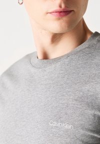 T-shirt en coton gris avec un col rond, arborant un petit logo "Calvin Klein" blanc sur la partie gauche de la poitrine. Texture lisse.