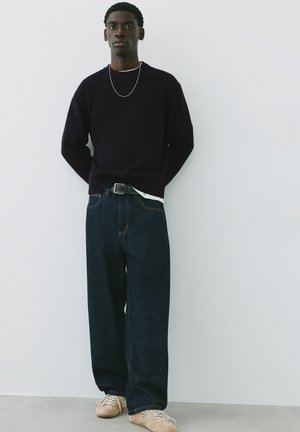 Mann steht an einer weißen Wand, trägt einen schwarzen Pullover, weite dunkle Jeans, beige Turnschuhe und eine dünne Kette.