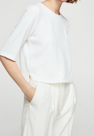 Vrouw draagt een getextureerd wit kort mouwtopje en losse witte broek met handen in de zakken tegen een effen achtergrond.