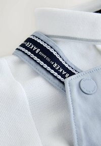 Gros plan sur un tissu blanc texturé avec une bordure bleu clair, un bouton marqué "Baker by Ted Baker", et une sangle marine avec le texte "Baker by Ted Baker".