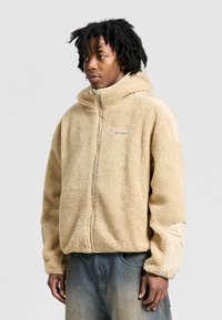 Beige fleecejacka med dragkedja fram, huva och kontrasterande släta ärmar. Har en avslappnad passform och texturerad yta.