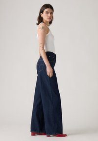 Temno modre kavbojke širokih nog z visokim pasom, z zadnjim žepom z vezenim logotipom, kombinirane z belo majico brez rokavov in rdečimi čevlji.