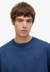 Sudadera azul marino de tela suave, con cuello redondo y detalle de logo bordado en el lado izquierdo del pecho, con una textura lisa y uniforme.