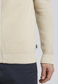 Beige geribbelde trui met mouwen en zoom, gedragen door een persoon in donkerblauwe broek met een zichtbare hand aan de zijkant.