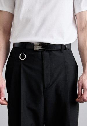 Curea din piele neagră cu o cataramă argintie strălucitoare, având un detaliu suplimentar sub formă de inel circular. Asortată cu pantaloni negri și o cămașă albă.