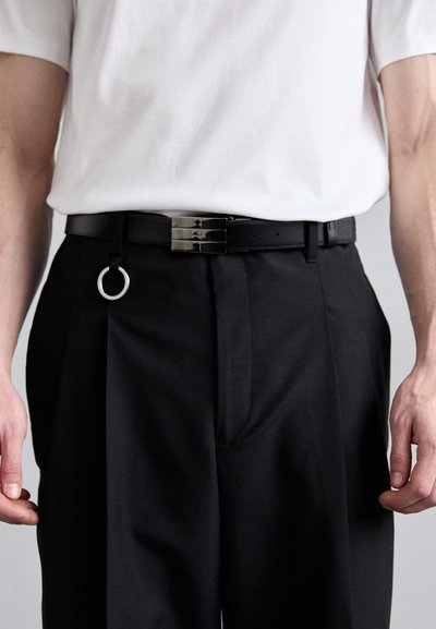 Schwarzer Leder-Gürtel mit einer glänzenden silbernen Schnalle, die ein zusätzliches kreisförmiges Ringdetail aufweist. Kombiniert mit schwarzen Hosen und einem weißen Hemd.