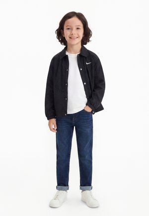 Lachend kind met middellang donker haar, draagt een zwart jasje over een wit hemd, blauwe opgerolde spijkerbroek en witte sneakers, staat met hand in de zak.