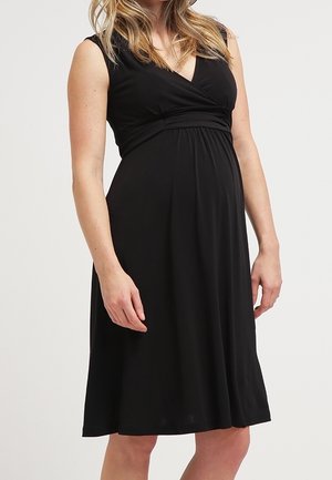 Schwangere Frau trägt ein ärmelloses schwarzes knielanges Kleid mit tailliertem Schnitt und V-Ausschnitt, steht vor einfarbigem Hintergrund.