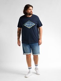 Tmavěmodré tričko s diamantovým motivem a nápisem "Petrol Ind." v kombinaci se světle modrými džínovými šortkami a bílými teniskami s černými pruhy.