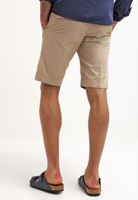 Homme portant un short beige aux genoux, une chemise bleu marine et des sandales noires à enfiler, debout les mains dans les poches, tourné le dos.