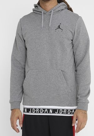 Mand iført en grå Jordan hættetrøje med sort Jumpman-logo på brystet og sorte shorts med rød stribe og Jordan-logo i livremmen.