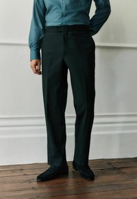 Tmavozelené priliehavé nohavice s hladkou textúrou, plochým predným dielom a jemnými záhybmi, doplnené čiernymi nubukovými topánkami.