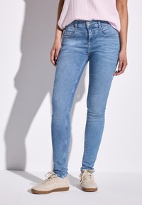 Jean skinny bleu clair avec une texture lisse, cinq poches et une fermeture à boutons, associés à des baskets beiges.