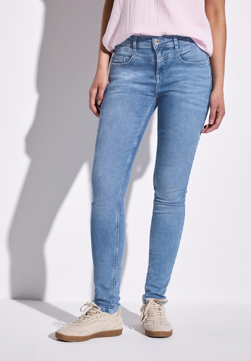 Jean skinny bleu clair avec une texture lisse, cinq poches et une fermeture à boutons, associés à des baskets beiges.