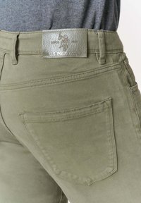 Pantaloni verde oliva con una toppa in pelle con logo. Presentano tasche posteriori, dettagli cuciti e elementi in metallo.