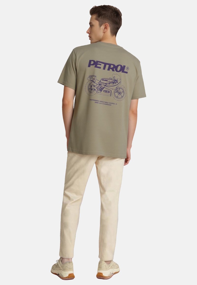 Oliwkowa koszulka z grafiką motocykla w granatowym kolorze na plecach. Połączona z jasnobeżowymi spodniami i neutralnymi sneakerami.