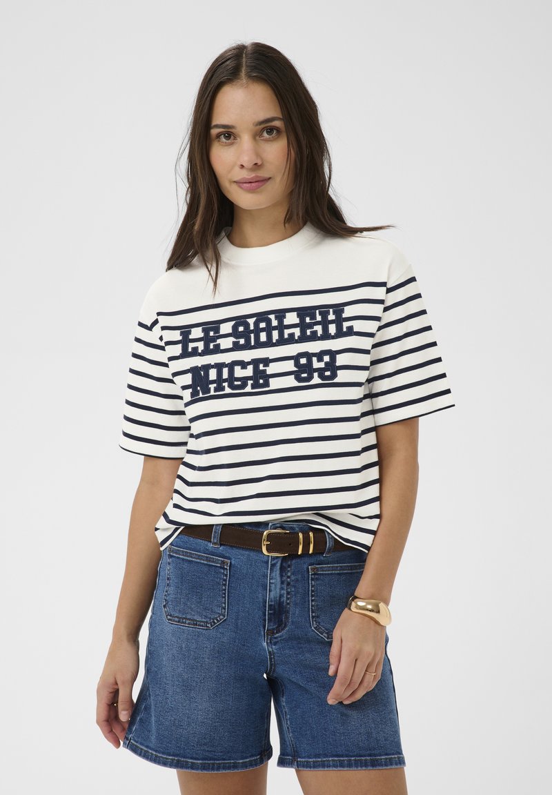 Vrouw draagt een zwart-wit gestreept T-shirt met de tekst "LE SOLEIL NICE 93", blauwe denimshorts, een bruine riem en een gouden armband.
