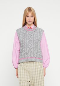 Szary dzianinowy bezrękawnik z warkoczowym wzorem i różowym akcentem na dole, noszony na różowej koszuli z kołnierzykiem i zestawiony z jasnymi, kraciastymi spodniami.