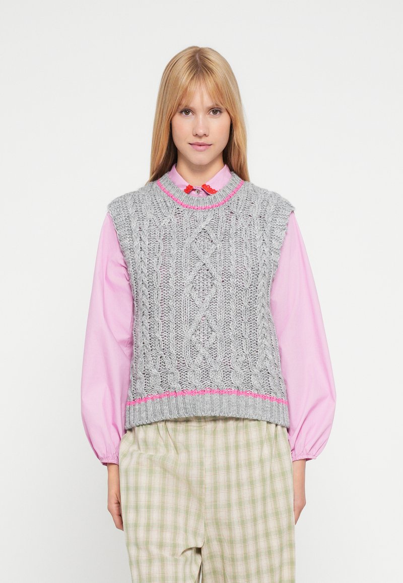 Szary dzianinowy bezrękawnik z warkoczowym wzorem i różowym akcentem na dole, noszony na różowej koszuli z kołnierzykiem i zestawiony z jasnymi, kraciastymi spodniami.